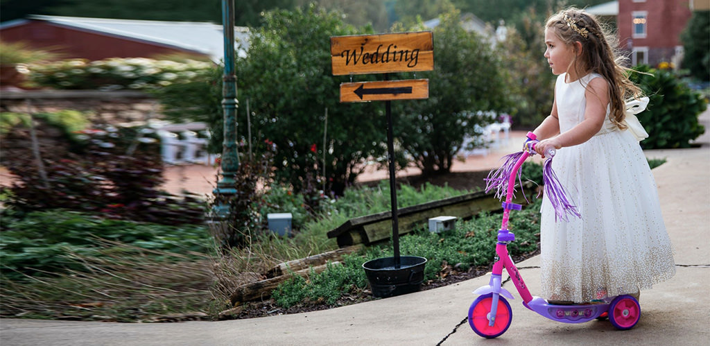 A cute flower girl riding down the aisle on a pink and purple scooter. This is for the article called non-traditional wedding ceremony ideas